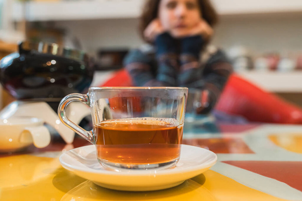 Drinking tea in tea room. filled cup of tea on table. Tea cafe, cozy place to spend your leisure time, warm up with hot drink in cold weather. Teen girl in warm sweater in background out of focus - Photo, Image