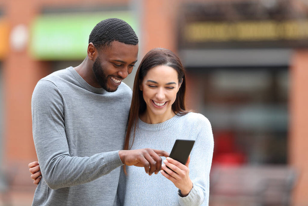 Feliz pareja interracial revisando el teléfono celular riendo al aire libre en la calle - Foto, imagen