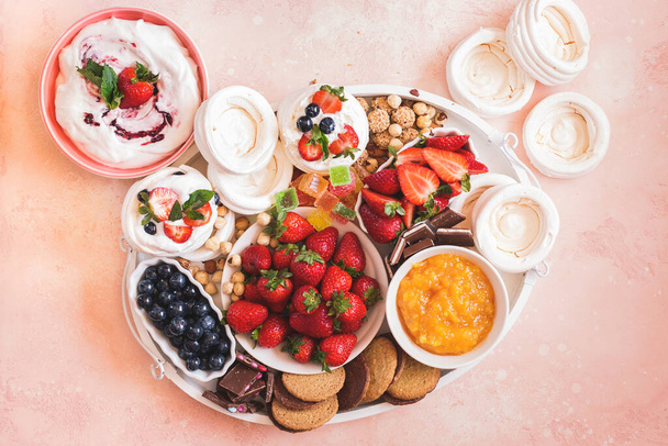Mini meringue berry sweet dessert platter. Small nests of meringue  on wooden tray with fresh fruits, ginger cookies, toppings, chocolate and whipped cream. Top view, blank space - Foto, Imagen