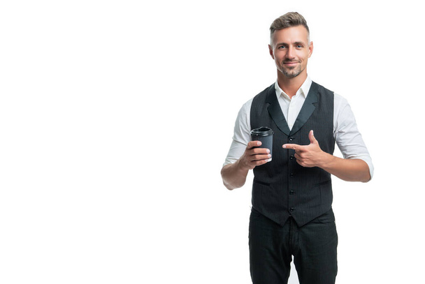 Szczęśliwy facet w kamizelce garnituru wskazując palcem na jednorazowy kubek z herbatą na wynos izolowane na białym - Zdjęcie, obraz