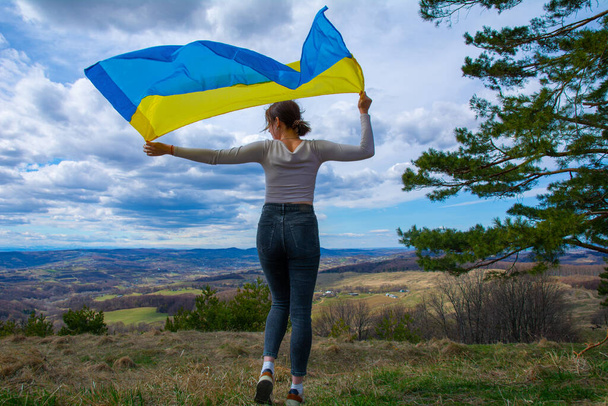 Una mujer joven con una bandera de Ucrania en el fondo de la hermosa vista a las montañas y la naturaleza ucraniana y el cielo nublado dramático. Mujer patriótica con bandera ucraniana. - Foto, imagen