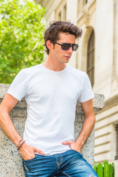 Man Summer Casual Street Fashion. Wearing White T shit, jeans, sunglasses, hands in pockets, a young European college student standing at corner on campus in New York, looking down, sad, thinking. - Foto, Imagen