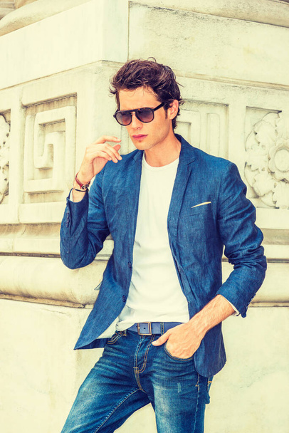 Ανδρική μόδα. Ντυμένος με μπλε σακάκι, λευκό κάτω από το πουκάμισο, τζιν, φορώντας γυαλιά ηλίου, ένας νεαρός Ευρωπαίος φοιτητής που στέκεται στην πανεπιστημιούπολη της Νέας Υόρκης, το χέρι σταματά στον αέρα, σκέφτεται. - Φωτογραφία, εικόνα