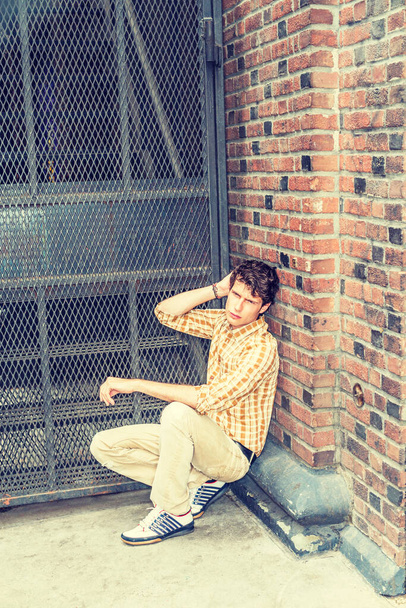 Concept of lonely man thinking about love. Wearing yellow patterned shirt, pants, sneakers, a young European guy squatting at corner on street in New York, sad, thinking after busy working day - Photo, Image