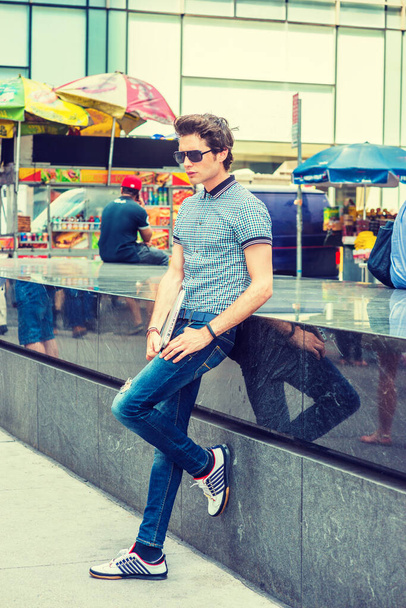 Студент Европейского колледжа в Нью-Йорке. Носит рубашку с короткими рукавами, джинсы, кроссовки, солнцезащитные очки, держит ноутбук, молодой парень, стоящий напротив мраморной стены скамейки на улице, думая: - Фото, изображение