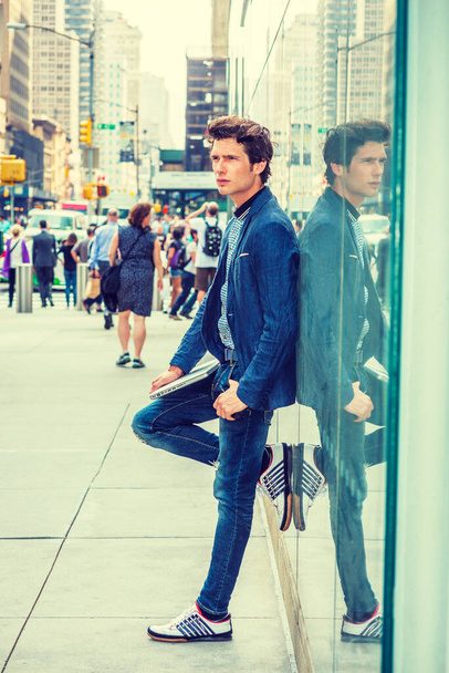 European College Student in New York. In blauem Blazer, Jeans, Turnschuhen, Laptop in der Hand steht ein junger Mann an einer Glaswand auf der Straße. Viele Menschen gehen im Hintergrund. - Foto, Bild