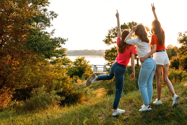 サマーキャンプ、サマークラス、フレンズ大学。3人の陽気な女の子の友達は、夏の日没の自然を背景に踊りジャンプします。夏休みに友人のグループ - 写真・画像