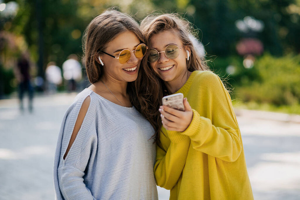 Girls outside in bright clothes and sunglasses in headphones, holding smartphone. Video call, online time with friends, outdoor time, warm sunny day in city. High quality photo - Fotó, kép
