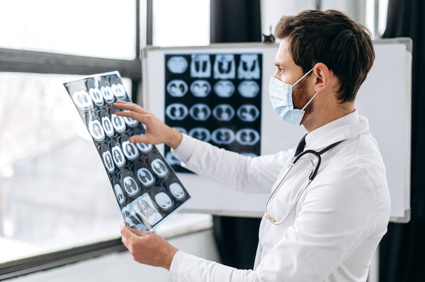 Slimme professionele blanke arts in een medisch uniform, beschermend masker, een pulmonoloog, een therapeut, staat in het ziekenhuis, onderzoekt een röntgenfoto van zijn patiënten longen, denkt na over het voorschrijven van de behandeling - Foto, afbeelding