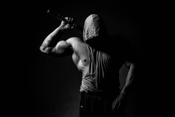 Dangerous muscular man in the mask with baseball bat ready for fight. An aggressive hooded man is holding a baseball bat on black. - Foto, immagini