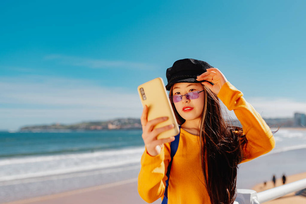 young asian woman takes a picture of herself with her mobile phone while sightseeing on a city beach. chinese girl taking a selfie - Zdjęcie, obraz