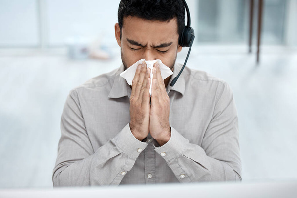 Got the sniffles. Shot of a young businessman blowing his nose while working in a call centre. - 写真・画像