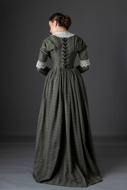 Una mujer victoriana de clase trabajadora con un corpiño y una falda de color verde oscuro con un cuello de encaje y de pie sobre un fondo gris del estudio - Foto, Imagen