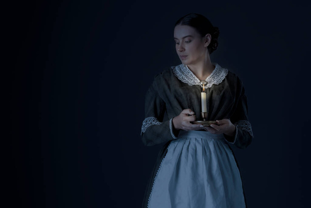 A maid servant or working class Victorian woman wearing a dark green checked bodice and skirt and holding a candle - Foto, Imagem