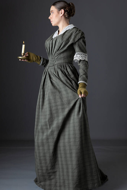 A maid servant or working class Victorian woman wearing a dark green checked bodice and skirt and holding a candle - Photo, Image