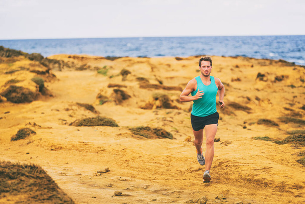 Running man male runner athlete cross country running on golden sand coast by ocean. Young men jogging training on trail outdoors - 写真・画像