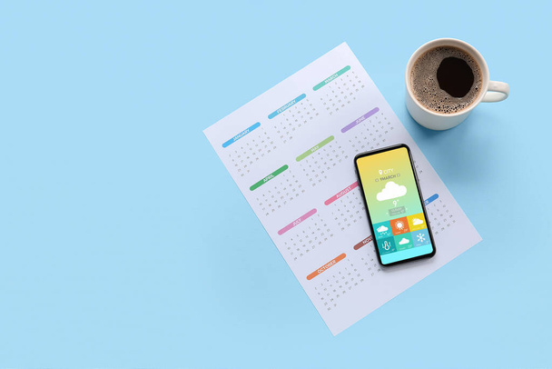 Calendário de papel, telefone celular com aplicativo de previsão do tempo e xícara de café no fundo azul claro - Foto, Imagem