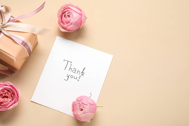 Sheet of paper with text THANK YOU, gift box and flowers on beige background - Фото, зображення