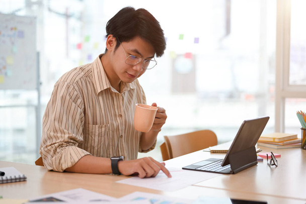 Focado empresário asiático ou empresário masculino tomando café enquanto verifica venda e relatório de estoque no escritório. - Foto, Imagem