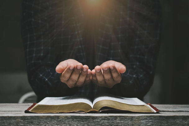 Man hands praying to god with the bible. Yellow lights and sparkles coming on Believe in goodness. Holding hands in prayer on a wooden table. Power of hope or love and devotion.  - Photo, Image