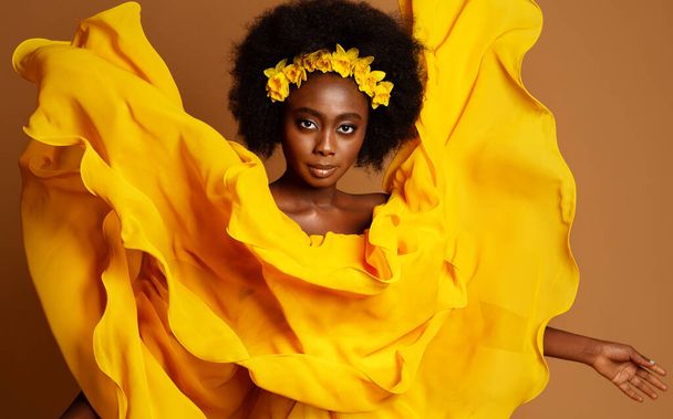 Dark Skinned Woman Fashion Portrait in Yellow Flying Dress. Beauty African Model with Flower Wreath in Black Afro Hairstyle. Dancer with fluttering Silk Fabric over Studio beige background - Foto, immagini