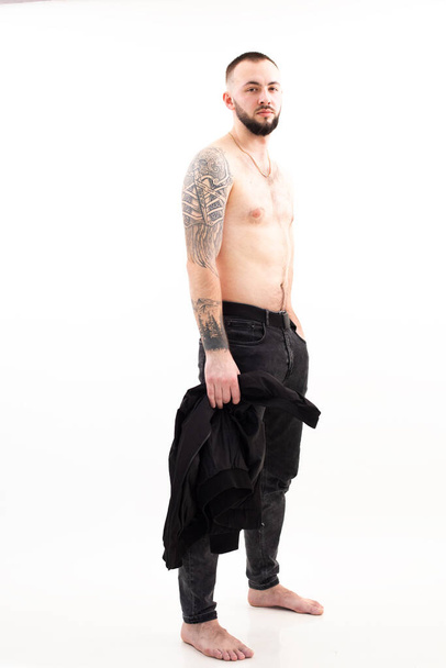 Portrait of young gorgeous bearded man with short dark hair wearing black jeans, golden chain with bare torso standing. - Foto, Imagen
