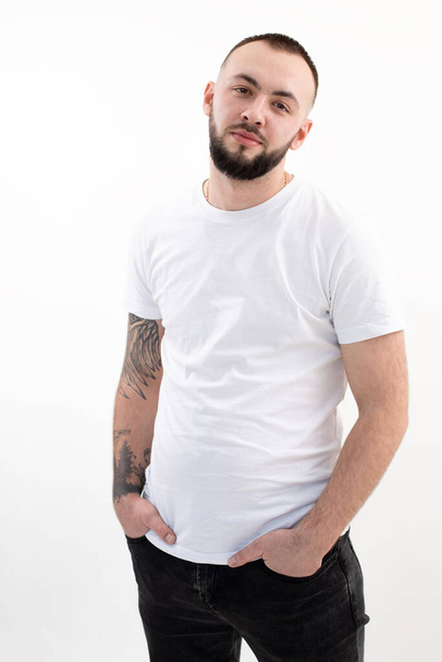 Young tranquil bearded man with short dark hair in white T-shirt, black jeans, stand thrusting hands into pockets. - Foto, Imagem