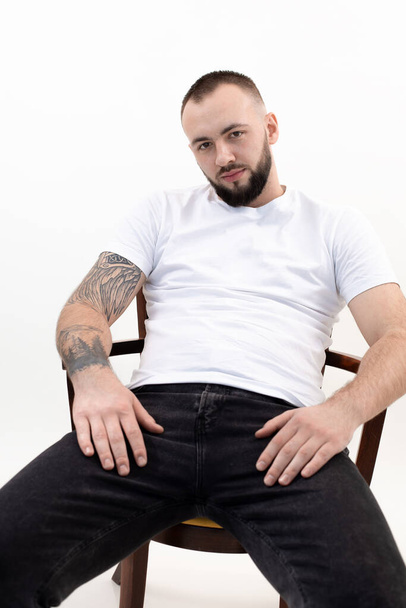 Young peaceful bearded man with short dark hair in white T-shirt, black jeans, sit on wooden chair, reclining at ease. - Foto, Imagen