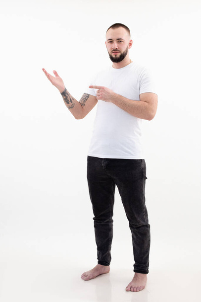 Portrait of young serious bearded man with short dark hair in white T-shirt, black jeans, stand pointing index finger. - Photo, Image