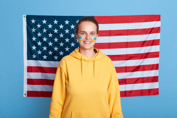 Indoor shot of positive woman wearing yellow hoodie and with flag of Ukraine on her cheek, isolated over blue background with USA flag, american support Ukraine during war against Russia. - Foto, afbeelding