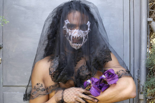 retrato de primer plano del joven sin camisa con máscara y velo negro sosteniendo orquídeas púrpuras - Foto, Imagen