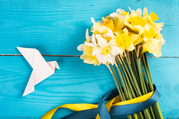 Yellow daffodils with a yellow-blue ribbon on a wooden background and a white dove made of paper. Origami. top view. Concept of Peace in Ukraine flat lay, lay out - Photo, image
