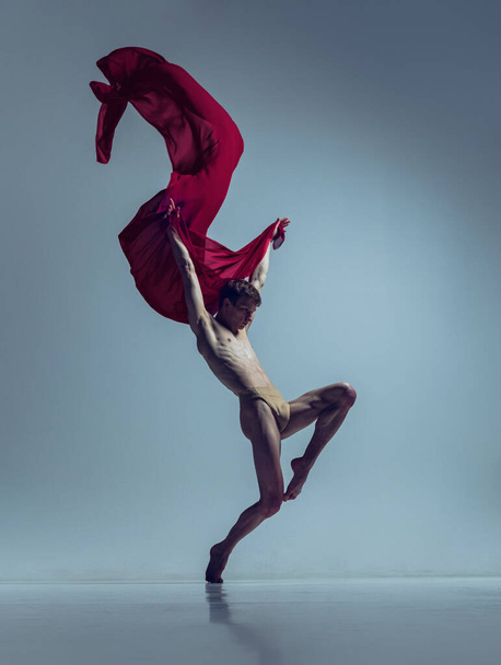 Portrait of young man, flexible ballet dancer in action with red fabric, cloth isolated on navy studio background. Grace, art, beauty concept. - Fotografie, Obrázek