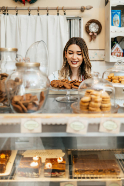Beautiful young female seller working in a confectionery shop or bakery. She is selling and packing delicious sweets. - 写真・画像