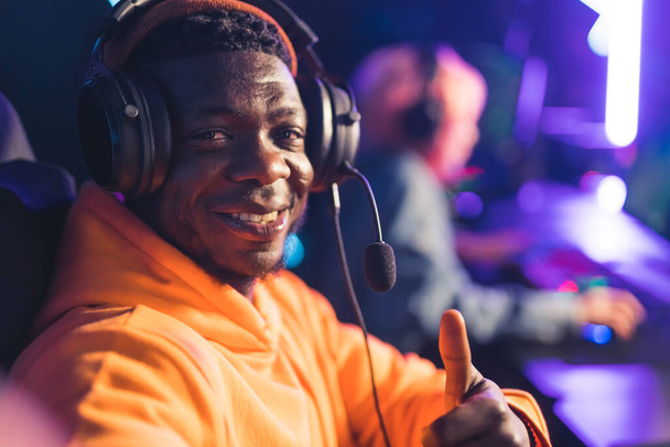 Smiling cheerful African-American male gamer with professional headphones gives a thumbs-up gesture to the camera. Gaming cafe interior. - Photo, Image