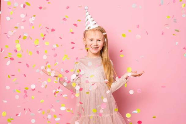 μικρό χαρούμενο νεαρό κορίτσι κάνει κόμμα με κομφετί στέκεται απομονωμένο πάνω από ροζ φόντο. - Φωτογραφία, εικόνα