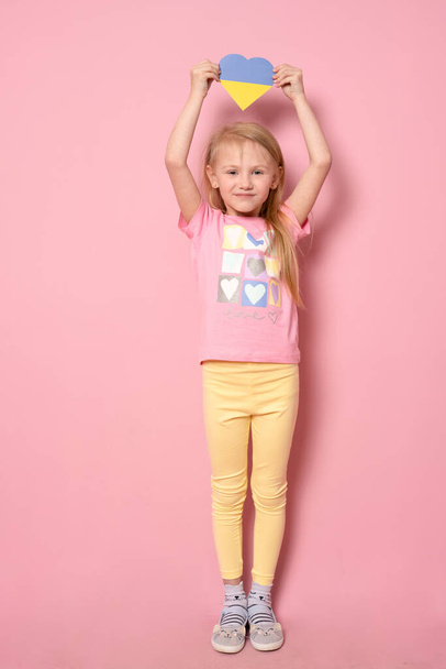 Portrait of a little girl standing on a pink background holding a heart in the colors of the flag of Ukraine. Children against war - Foto, imagen