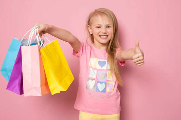 Photo of happy little caucasian girl standing isolated over pink background holding shopping bags and okay sign. Looking camera. - Фото, изображение