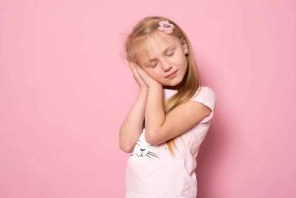 Relájate y duerme. Cansada linda niña caucásica de pie aislada sobre fondo rosa con los ojos cerrados apoyados en las palmas haciendo gesto de dormir. - Foto, Imagen
