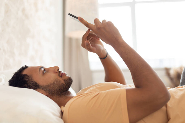 Arabic Man Using Smartphone Lying In Bed Texting In Bedroom - Foto, immagini