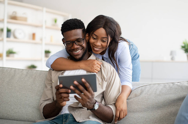 Fröhliches afroamerikanisches Paar sitzt mit Tablet auf Sofa - Foto, Bild