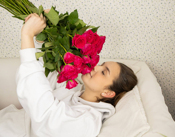 A young and happy girl lies on the bed and smells roses with her eyes closed - Фото, зображення