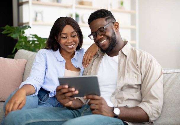 Cheerful African American couple sitting on sofa, using tablet - Fotoğraf, Görsel