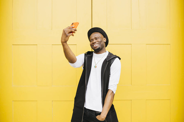 Portrait d'un jeune homme noir souriant prenant un selfie debout contre une porte jaune - Photo, image
