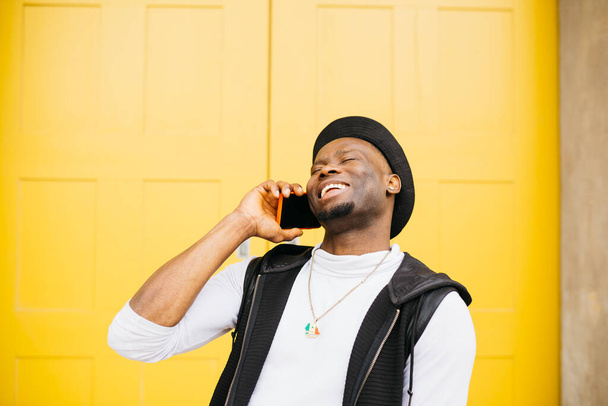Retrato de um jovem negro sorrindo falando em um telefone contra uma porta amarela - Foto, Imagem