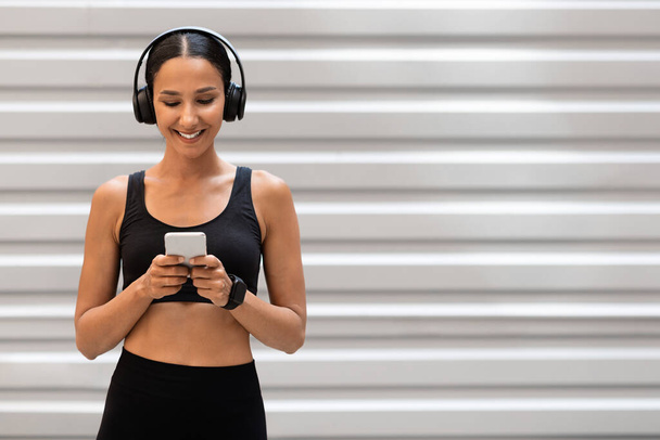 Sonriente joven árabe atleta femenina en ropa deportiva en auriculares charlando en el teléfono sobre fondo de pared gris - Foto, imagen
