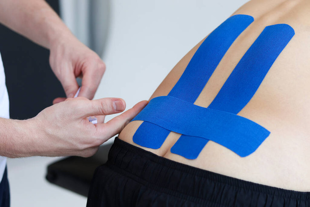 Физиотерапевт накладывает синюю ленту кинезио на нижнюю часть спины пациента. Кинезиология, физиотерапия, концепция реабилитации. - Фото, изображение