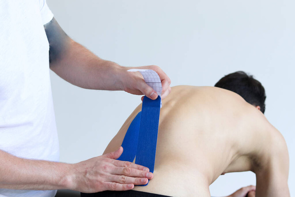Fysiotherapeut die blauwe kinesio tape op de onderrug van de mannelijke patiënt aanbrengt. Kinesiologie, fysiotherapie, revalidatieconcept. - Foto, afbeelding