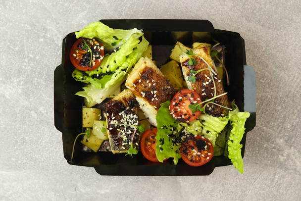 Healthy bowl with baked fish, potatoes, sesame, tomatoes and lettuce in eco paper container isolated on black background. Healthy food delivery service and daily ration concept. Close-up. - 写真・画像