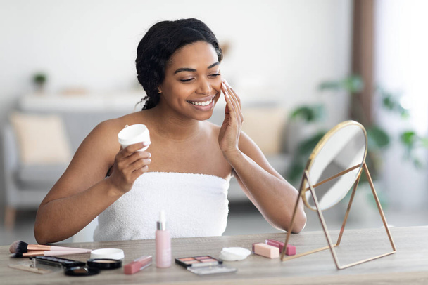 Cheerful attractive black woman using facial cream - Foto, Imagem